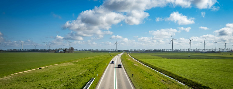 Panoramic image of a wind power plant on green farmland with cars driving on a crossing street on a sunny day